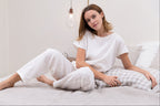Woman sitting on a bed wearing white lounge wear in a bedroom setting.