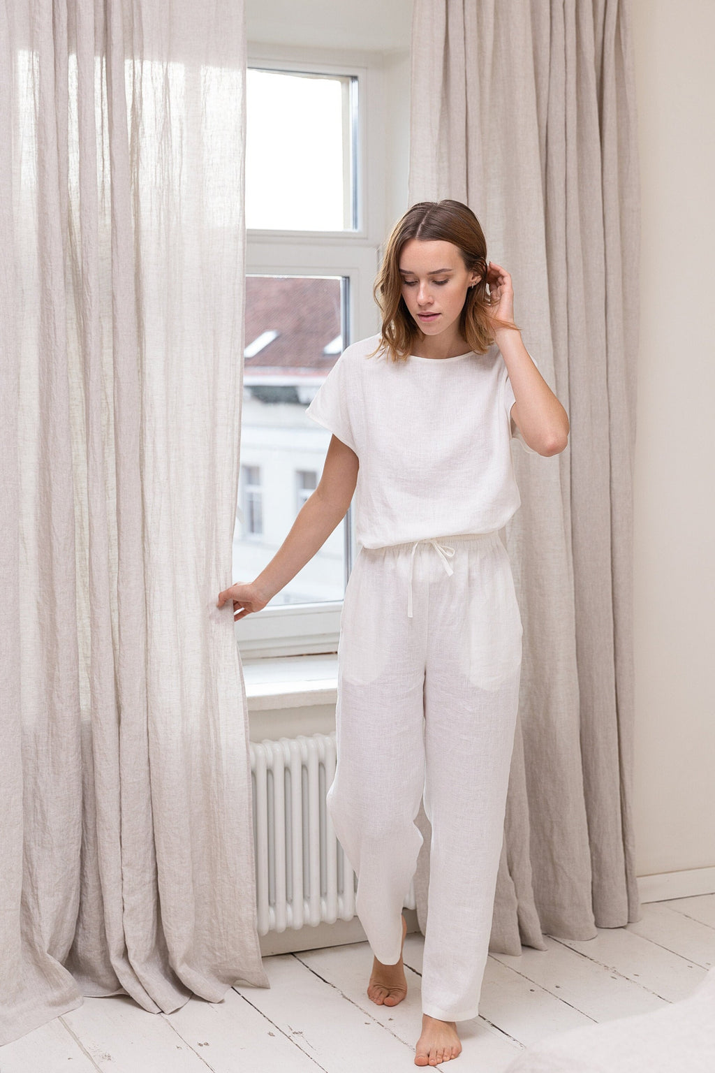 Woman in a white lounge set standing in a bright room with light-colored curtains.