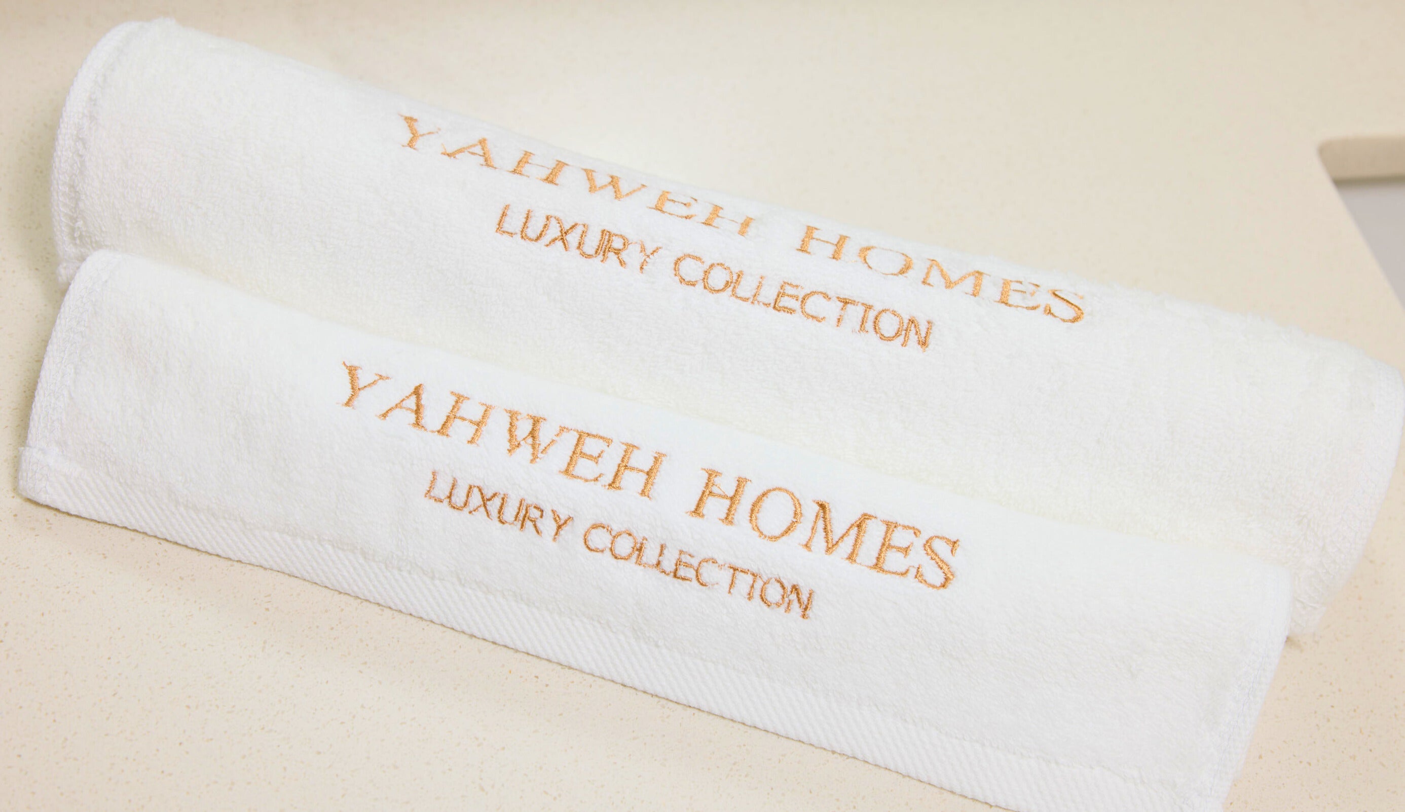 Two white towels with 'Yahweh Homes Luxury Collection' embroidery on a beige background